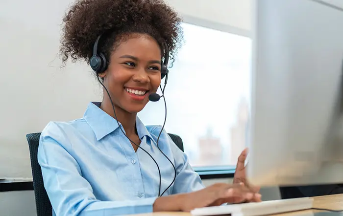 A smiling black woman in her twenties is sitting at a desk in front of a computer, wearing a telephone headset and speaking with a member