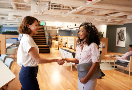 Two women shake hands in congratulations, one having landed a job and the other welcoming her to the team.