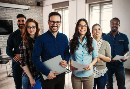 Six employees stand in a V-formation, smiling happily in their workplace, indicating job satisfaction and contentment.