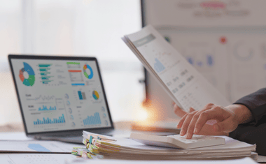 Bureau avec un ordinateur portable affichant des graphiques, des documents empilés et des mains tenant des feuilles.