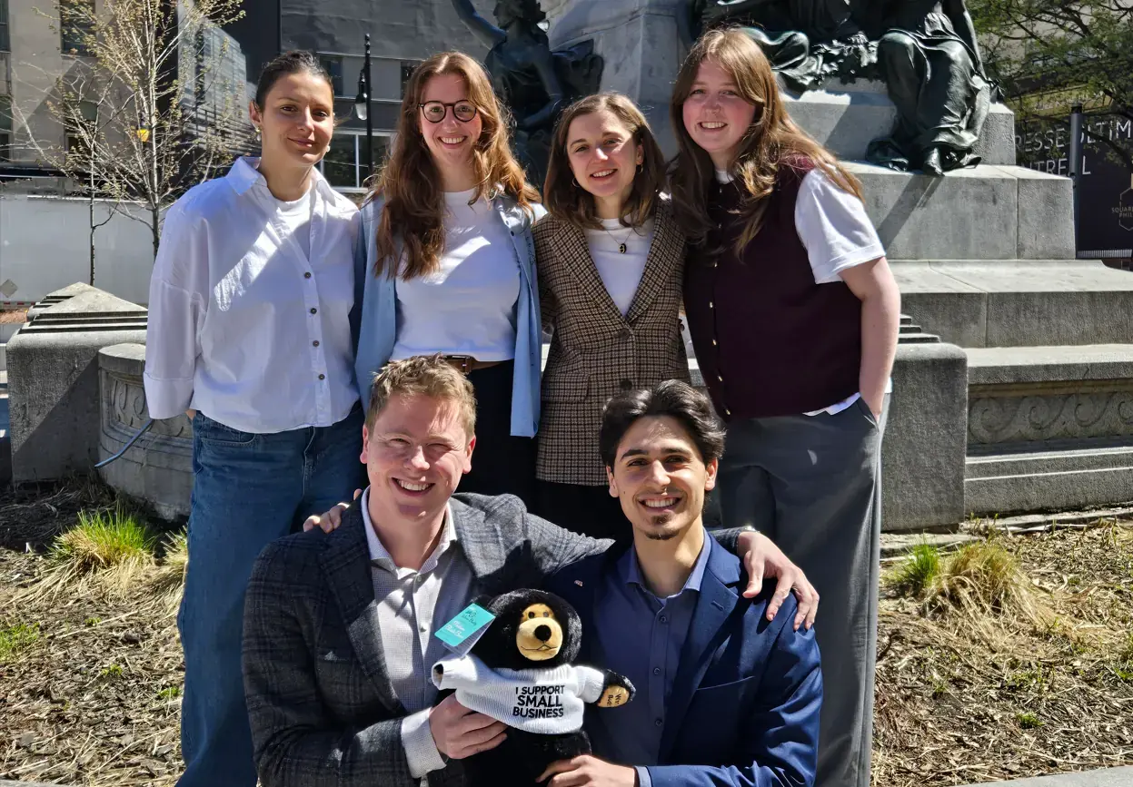 CFIB interns or former interns group photo outdoors in front of a monument, with several people standing and two seated in front holding a small plush toy wearing an I support small business shirt